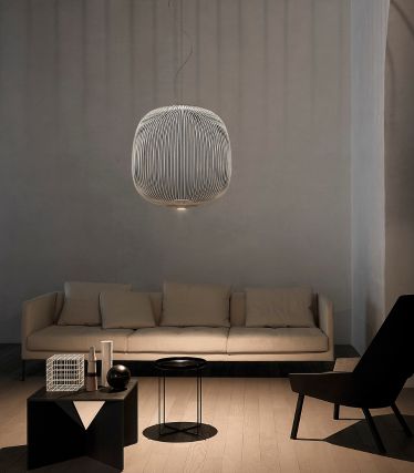 Un salon minimaliste avec la suspension Spokes de Foscarini comme point central élégant. Les délicates tiges métalliques de la lampe projettent de doux motifs lumineux sur les murs et le plafond, donnant à la pièce un effet sculptural et léger. Un canapé de couleur claire, des accents sombres et des matériaux naturels créent une atmosphère calme et moderne.