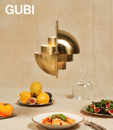 Une élégante suspension Multi-Lite de GUBI en laiton brossé est suspendue au-dessus d'une table dressée. Sous celle-ci se trouvent des assiettes de pâtes, des figues fraîches et des citrons, ainsi qu'une carafe en verre : une scène élégante qui allie design et plaisir.