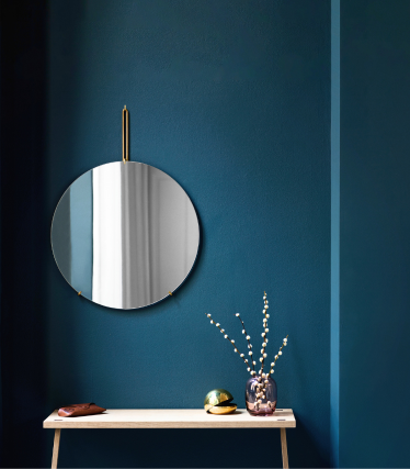 Un miroir mural rond avec une suspension en laiton est accroché à un mur bleu foncé. En dessous se trouve un banc en bois clair décoré d'un petit vase contenant des branches, d'un objet décoratif et d'un accessoire en cuir. La scène dégage une impression minimaliste, élégante et de style scandinave.