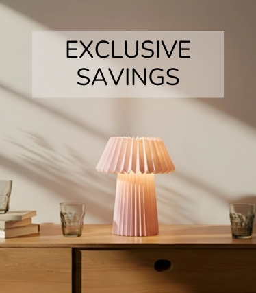 Lampe à poser rose pâle sur un buffet en bois, entourée de verres et de livres dans la lumière chaude du soleil.