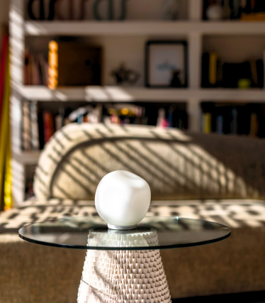 Petite lampe à poser blanche sur plateau en verre, lumière du soleil et bibliothèque en arrière-plan.