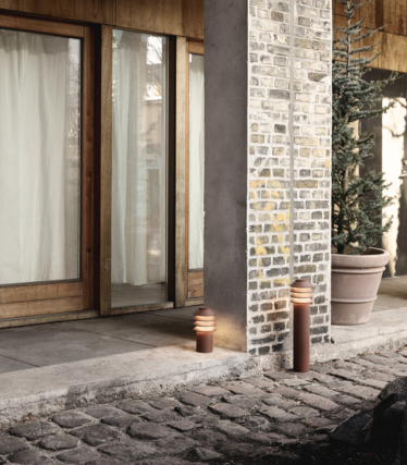 Scène extérieure avec deux lampadaires cylindriques modernes placés à côté d'une allée en pierre et d'une colonne en brique et béton. Derrière eux se trouvent des portes vitrées à cadre en bois avec des rideaux lumineux et une grande plante verte en pot. La lueur chaleureuse des lumières crée une atmosphère confortable et accueillante.