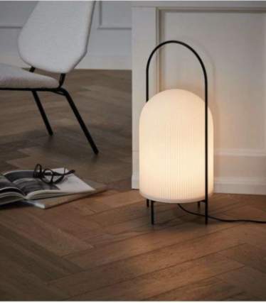 Lampe blanche cannelée avec une structure en arceau noir sur un parquet à chevrons, à côté d’un fauteuil.
